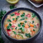 A bowl of chicken rice soup, shown with fresh parsley garnishes and lemon and bread on the background.