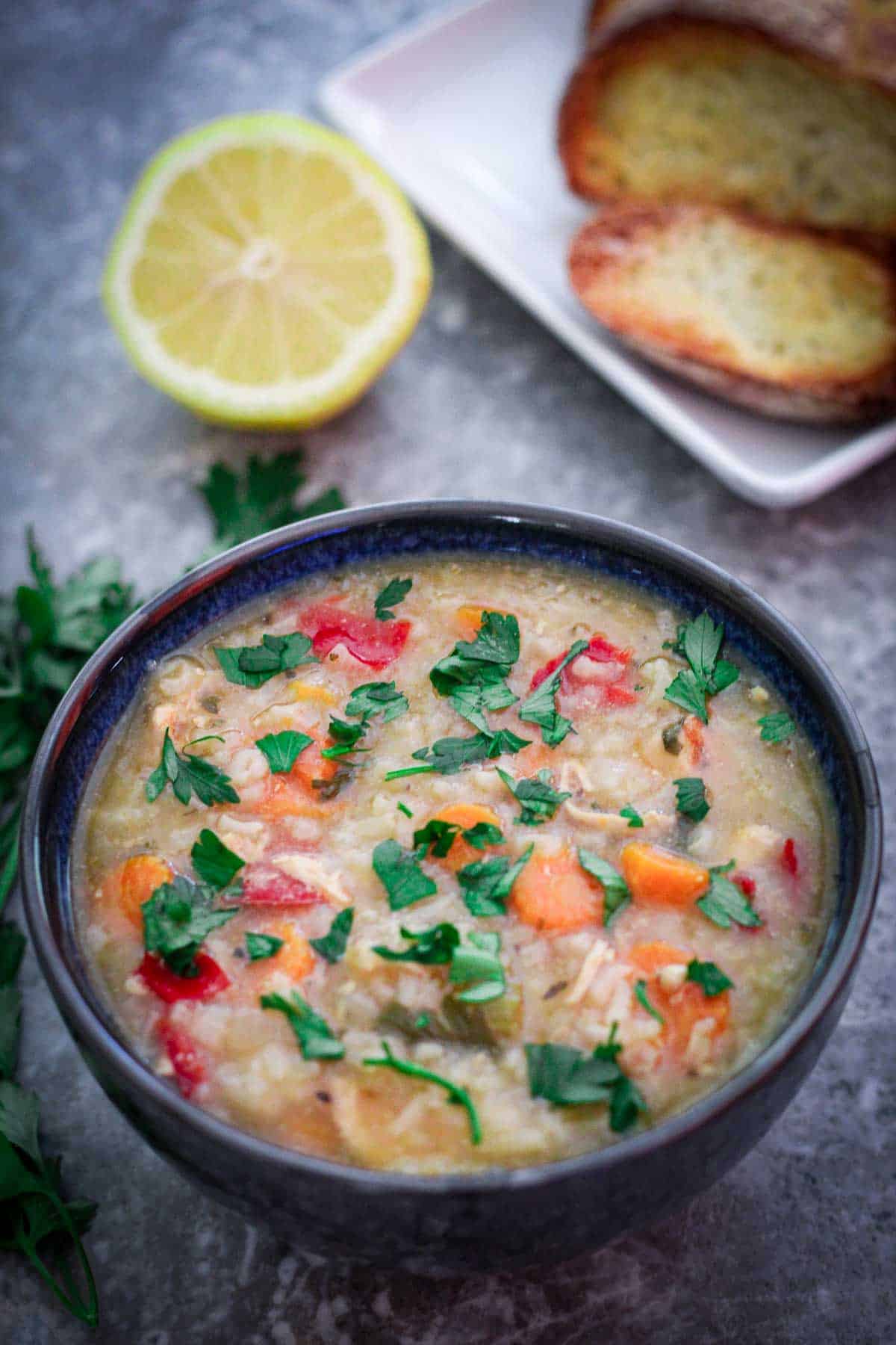 A bowl of chicken and rice soup, garnished with fresh parsley shown with a half lemon and toasted bread in the background.