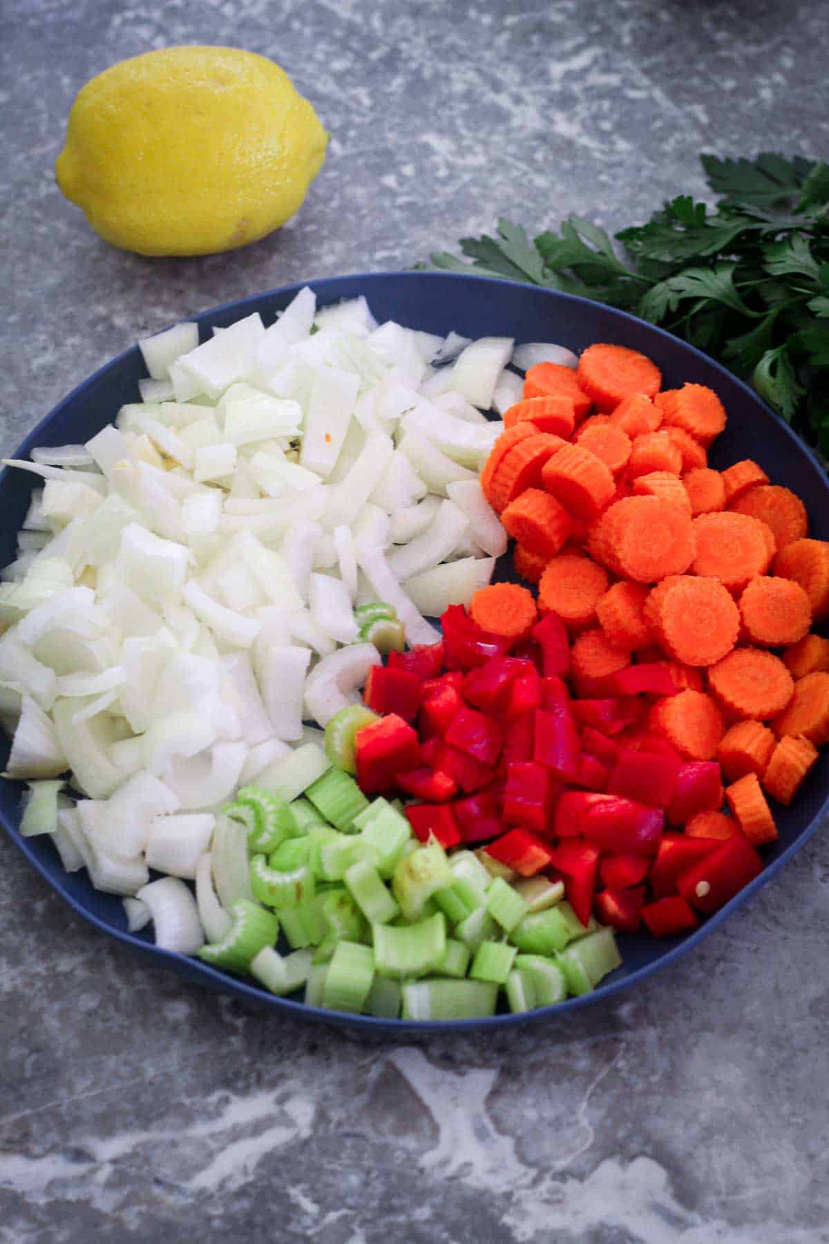 A plate with chopped onion, carrots, red bell pepper and celery. There's a lemon in the background and fresh Italian parsley.