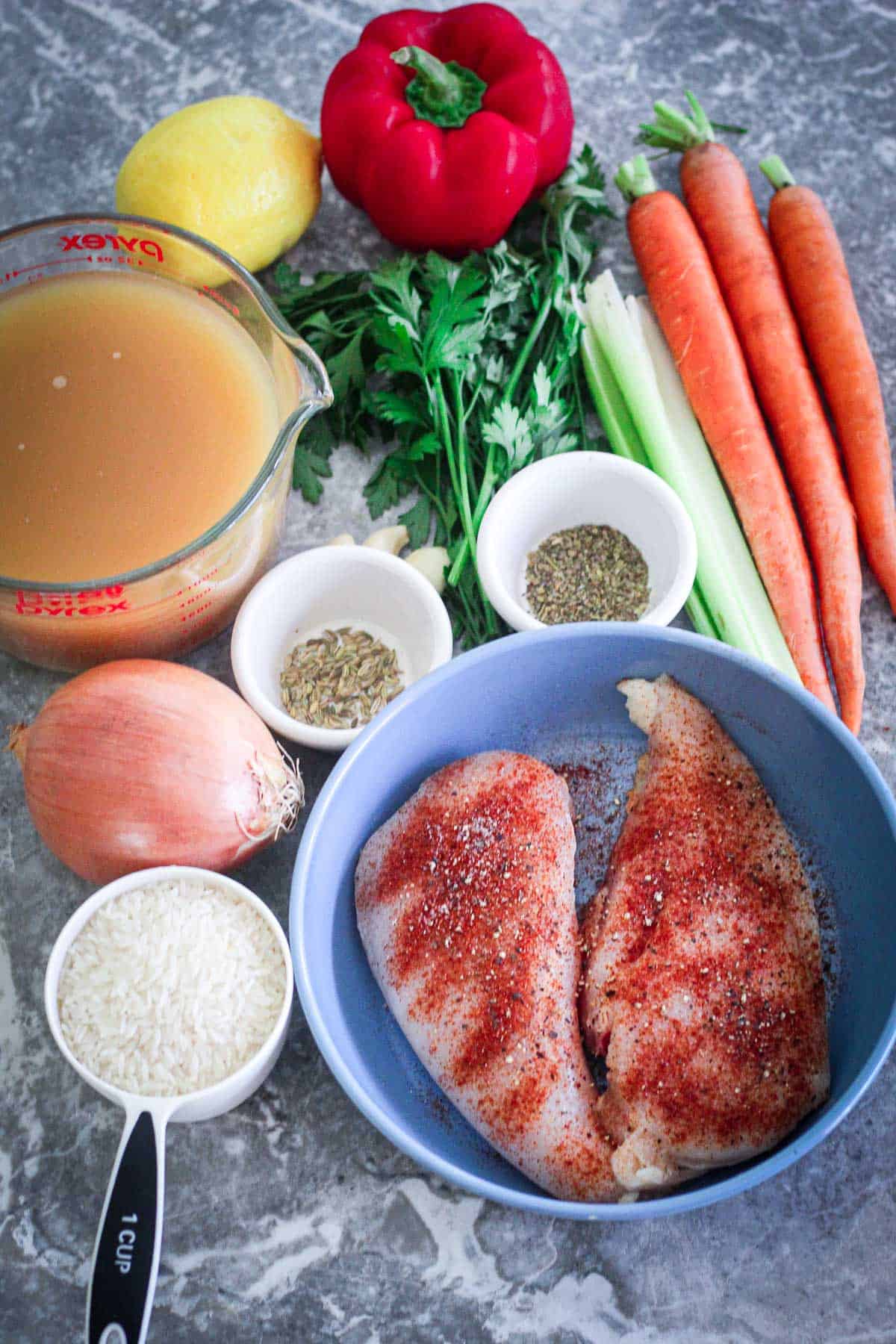 Ingredients for chicken and rice soup: broth, lemon, red pepper, carrots, celery, parsley, fennel seeds, Italian seasoning, yellow onion, rice and seasoned chicken breasts.