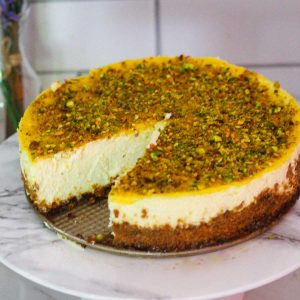 A cheesecake on a cake stand. Cheesecake has a bright yellow topping made with lemon curd and crushed pistachios over the curd. There's a slice of cheesecake already cut so you can see the remaining cheesecake and the triangular cut .