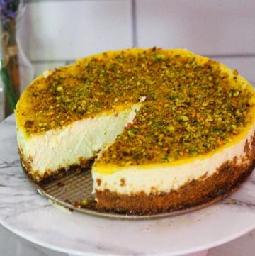 A lemon cheesecake with pistachios shown over a cake stand, a slice has already been cut out so you can look into in the cheesecake itself.