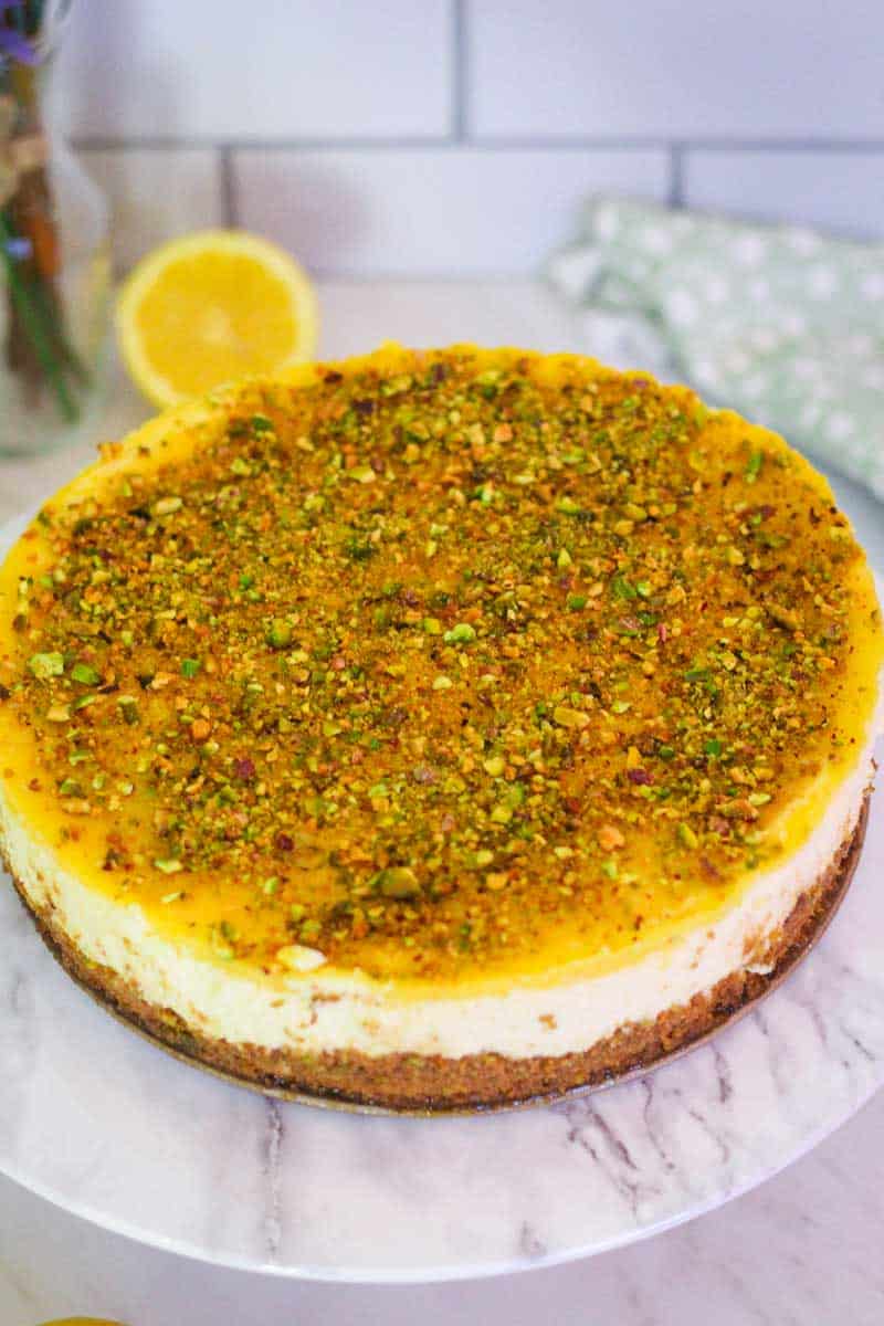 Lemon cheesecake, ready to slice on a cake stand. Cheesecake has a thin layer of lemon curd on top as well as crushed pistachios. 