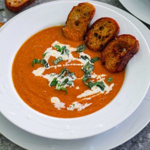 A soup plate over another plate, showing tomato soup with croutons dipped in it. Soup is garnished with basil and cream.