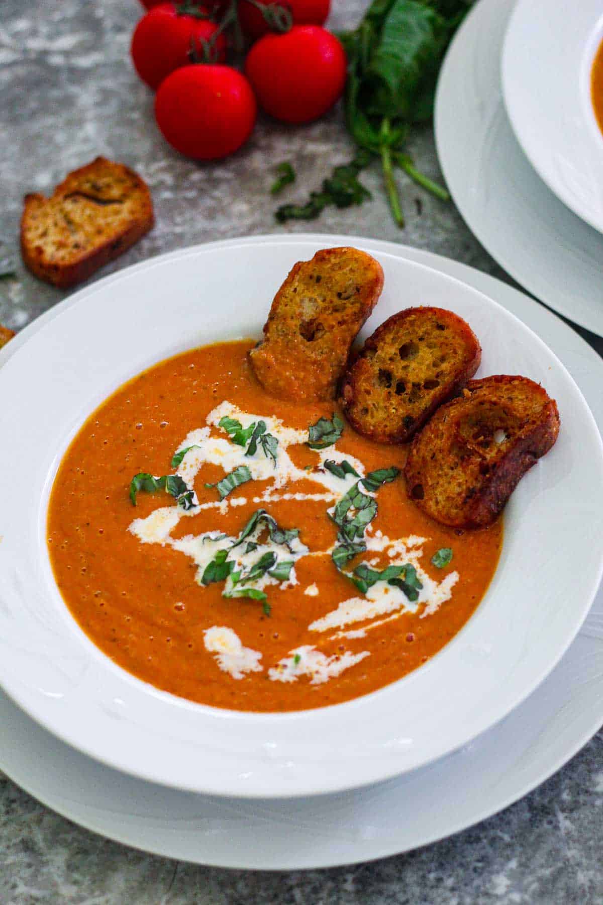 Roasted tomato soup shown served on a white plate and garnished with croutons, heavy cream and fresh basil.