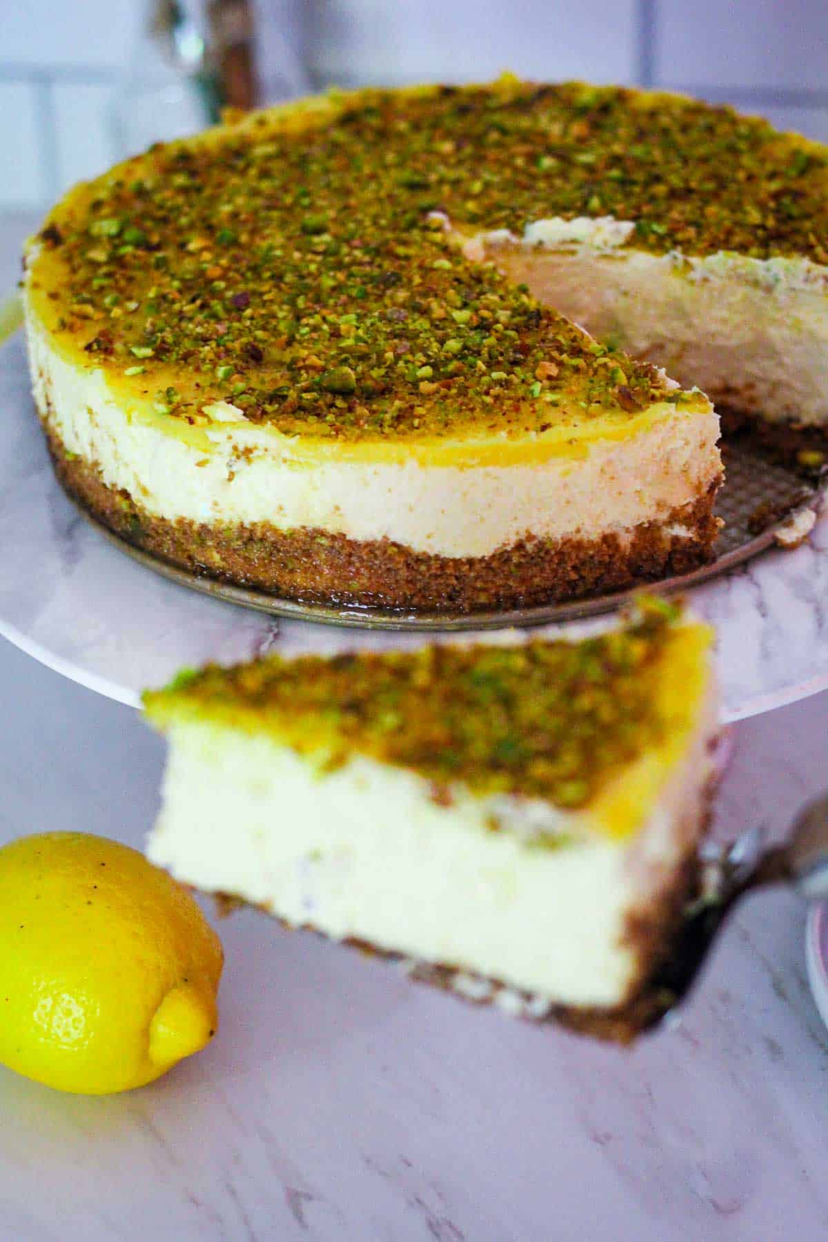 Cutting a slice of lemon cheesecake out of the cheesecake on a cake stand. Cheesecake is topped with lemon curd and pistachios. 