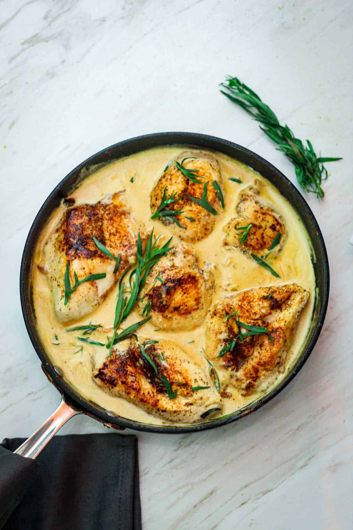 Skillet chicken with tarragon in a white wine sauce and dijon mustard. There's tarragon garnishes over the chicken and around the skillet.