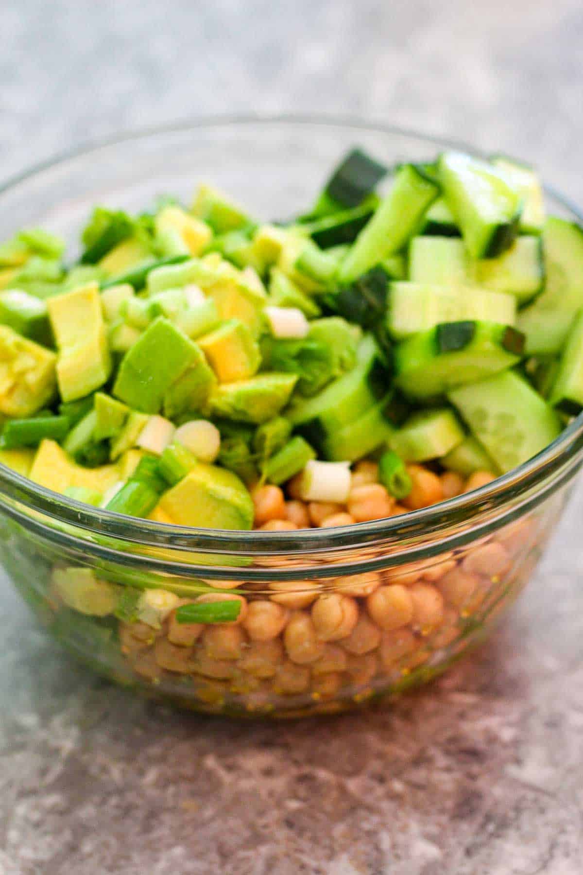 A medium glass bowl showing chickpeas, cucumbers, avocados, green onions etc ready to get tosssed.