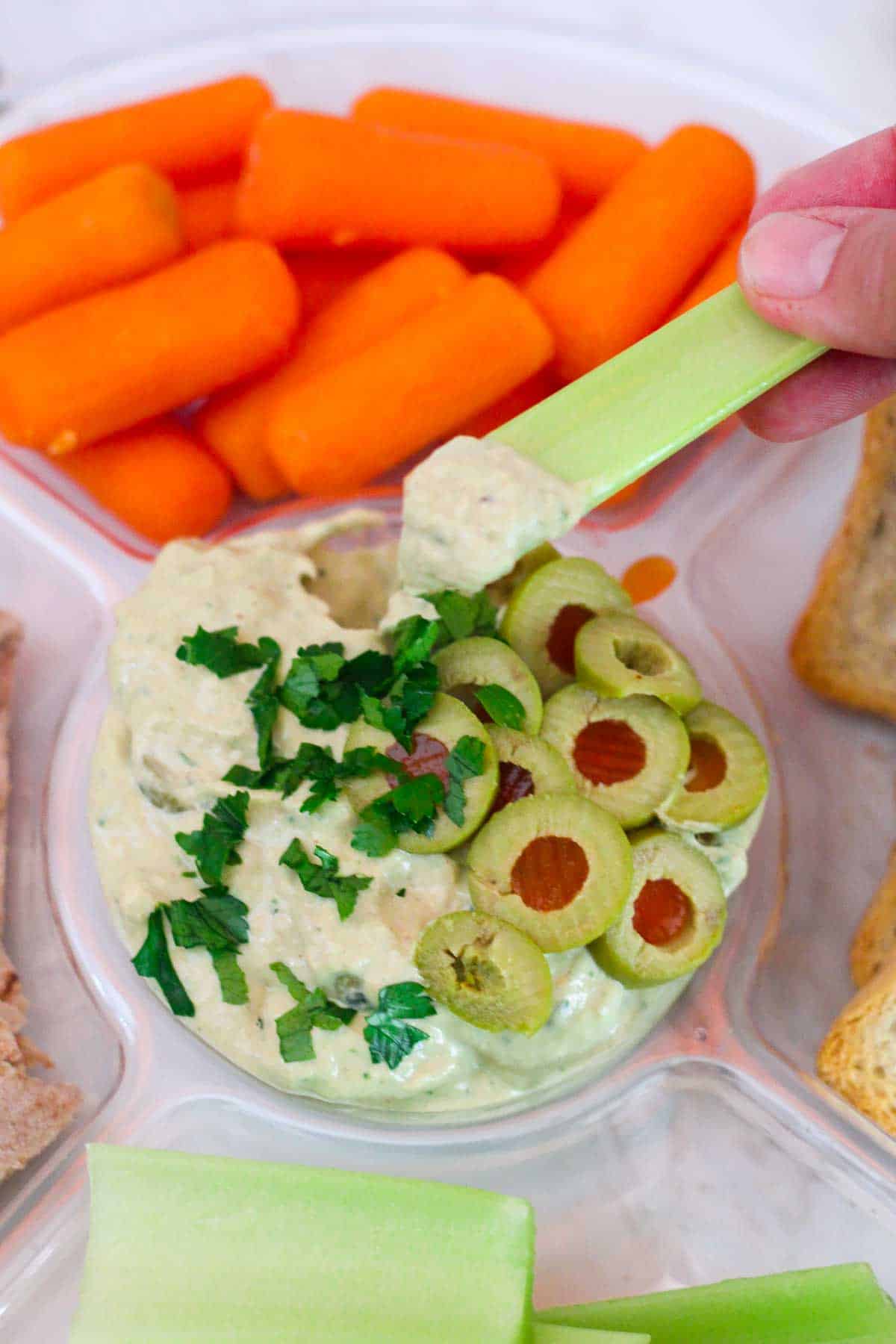Tuna dip garnished with sliced green olives and parsley. There's a celery stick being dipped in the tuna dip in this picture. You can see carrots, and other sides next to the dip.