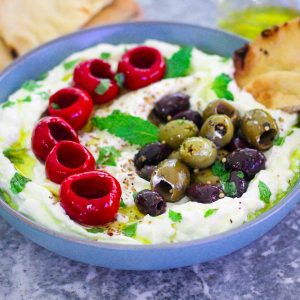 A round blue bowl with cottage cheese and feta dip, topped with olive oil, olives and marinated red peppers.