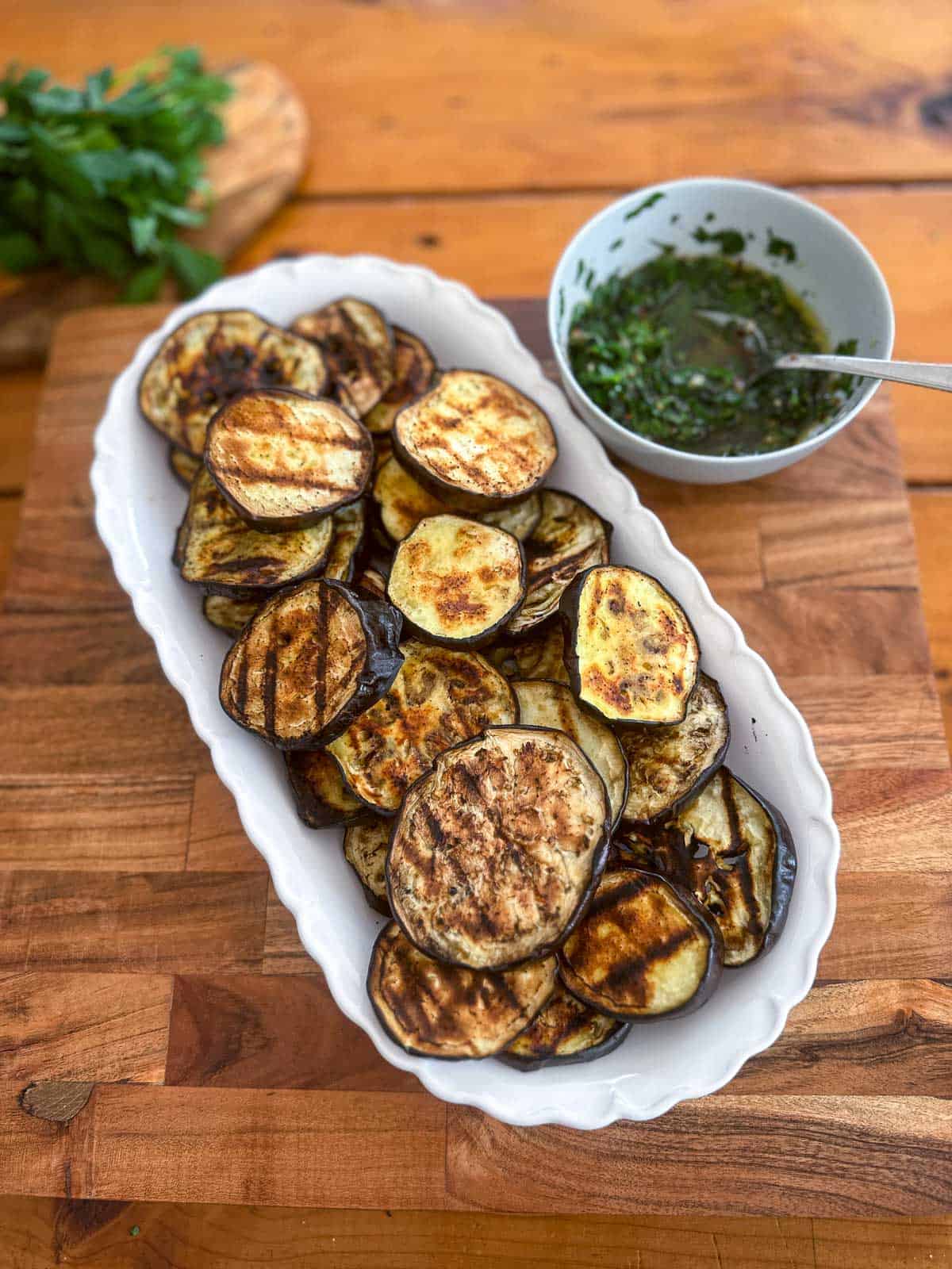 Grilled eggplant slices on a white tray with the parsley sauce next to it. sauce hasn't been poured on top yet so you see the grill marks on the eggplant clearly.