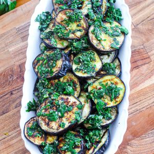A white serving tray showcasing grilled eggplant slices topped with a parsley and za'atar sauce!!