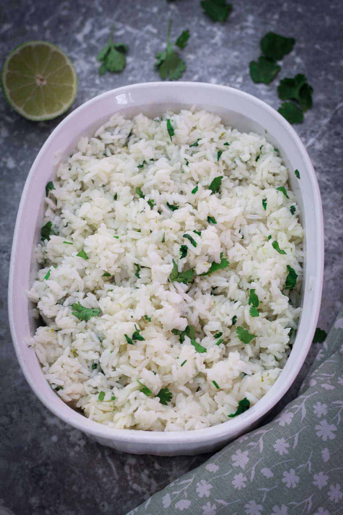 A white ceramic, oval shaped bowl showing cilantro, lime jasmine rice with garnishes around the bowl.