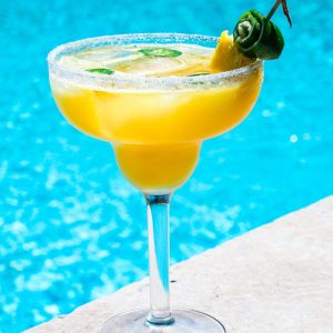A margarita glass on the edge of the pool showing pineapple margarita. Glass rim has salt and drink has a pineapple and lime garnish.