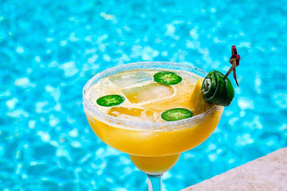 Poolside margarita showing a yellow drink with ice, with few slices of jalopeño floating over. Glass is garnished with a pineapple slice and lime arrangement. 