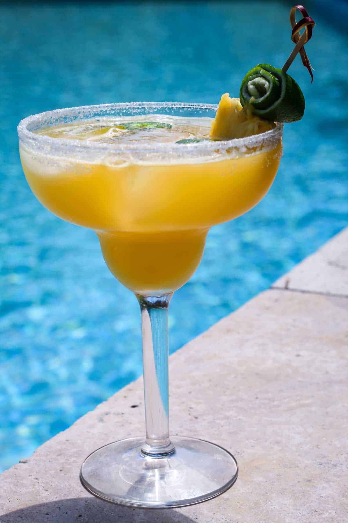 A glass with pineapple margarita, shown poolside with crystal blue water in the background. Drink is garnished with lime peel, pineapple slice and has floating jalapeño slices on top.