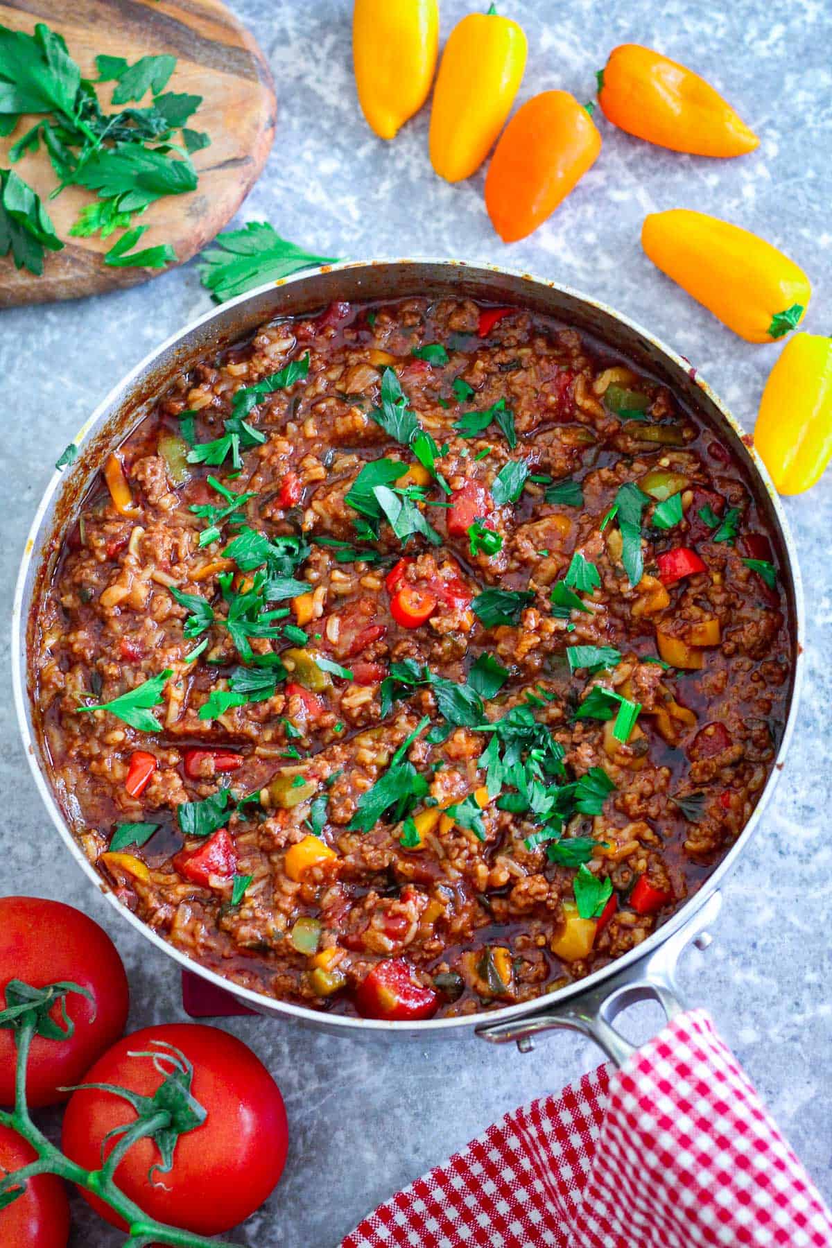 A skillet with ground beef, peppers, tomatoes and lots of seasonings and herbs shown in the middle. There's peppers, tomatoes, and parsley around the skillet.
