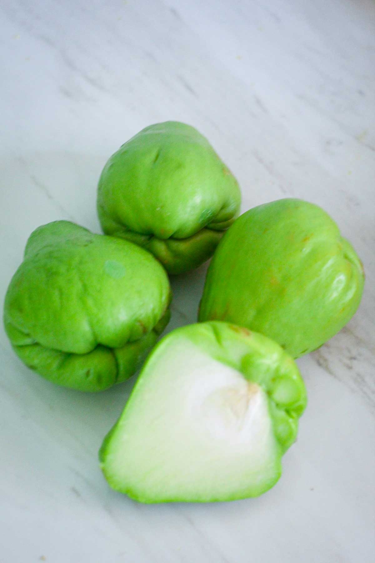 Four chayote squash on the counter, one is cut in half to show the interior of the fruit.