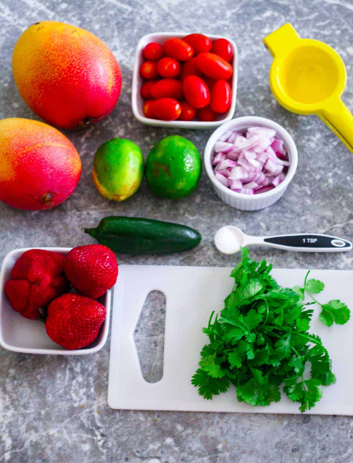 Ingredients for salsa lined up: 2 mangoes, 3 strawberries, a cup of grape tomatoes, chopped red onion, a jalapeño, a tso of seasalt, cilantro, limes and a citrus squeezer. 