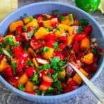 A round blue bowl with chopped mango strawberry salsa. There's a spoon sticking out and garnishes around the bowl.