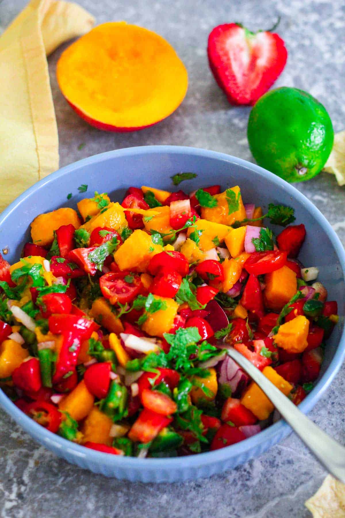 A colorful salsa with mango, strawberries, cilantro etc on a blue bowl surrounded by lime, mango and strawberry on the counter.