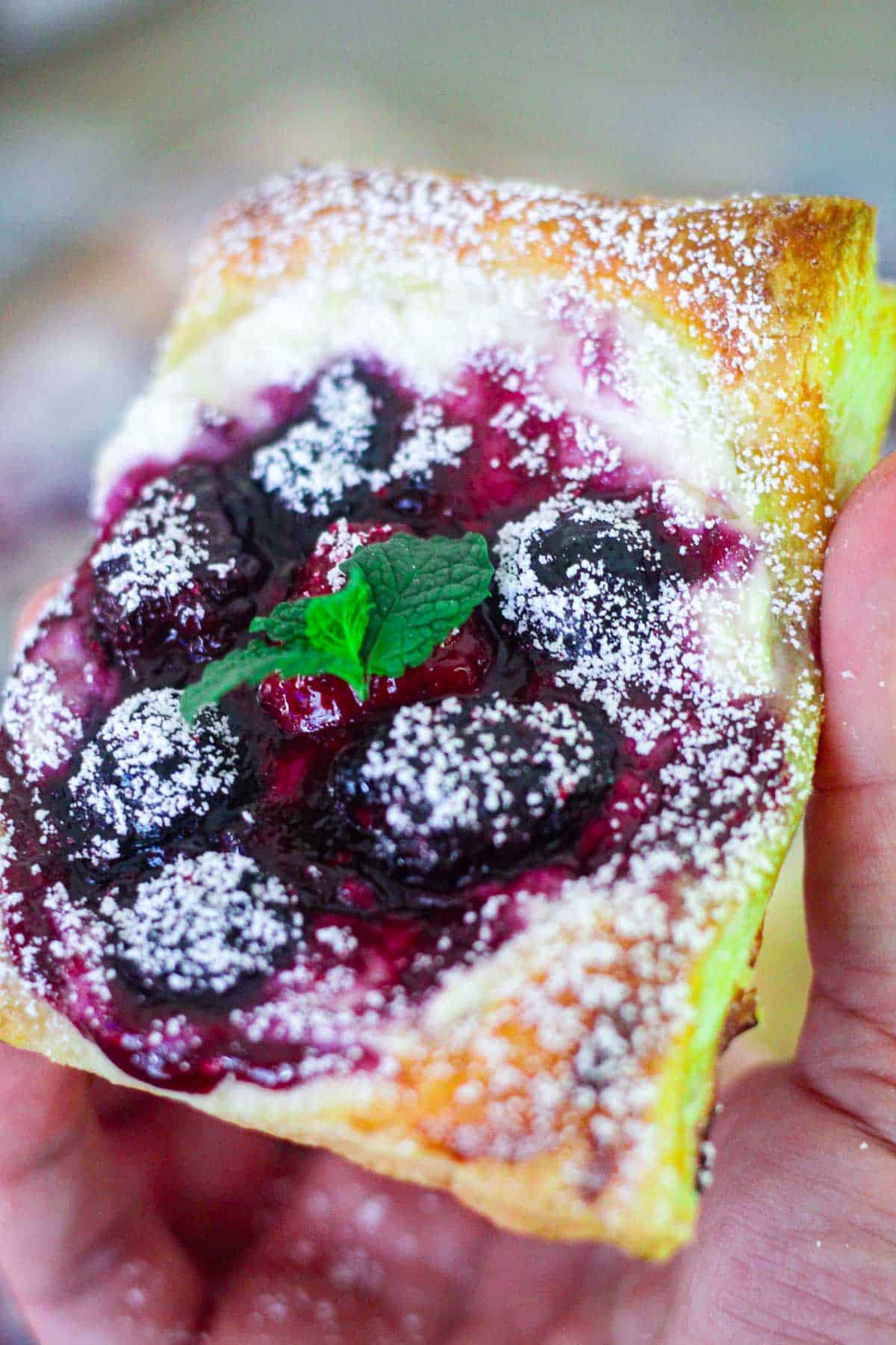 A single puff pastry tart with berries, shown towards the camera being held in one hand. Tart is garnished with powdered sugar and mint leaves.