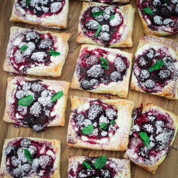 Puff pastry tarts with mixed berries and cottage cheese, sprinkled with powdered sugar are served on a wood board. They seem to be garnished with a little mint leaf individually.