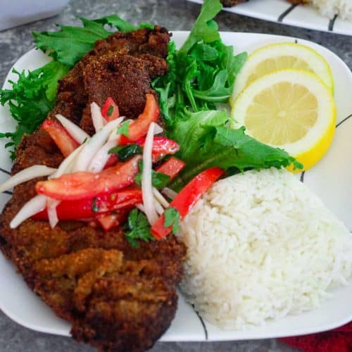 A dinner plate showing a breaded steak, topped with tomatoes and onions, next to rice and lemon slices.