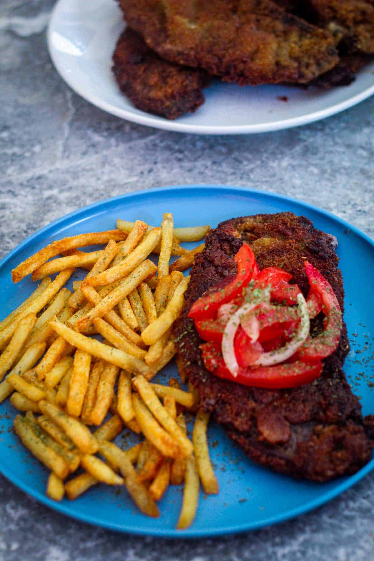 Milanesa steak served with french fries and topped with tomatoes, onions and garnished with some parsley.