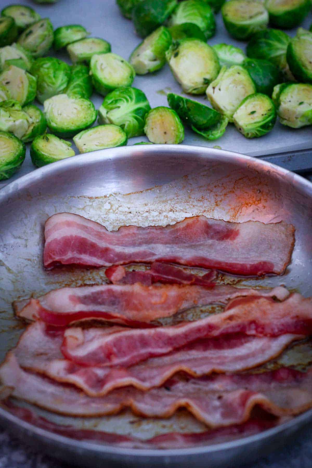 Partially seared bacon strips on a pan, next to the pan you see the roasting pan with the sprouts on it already. 