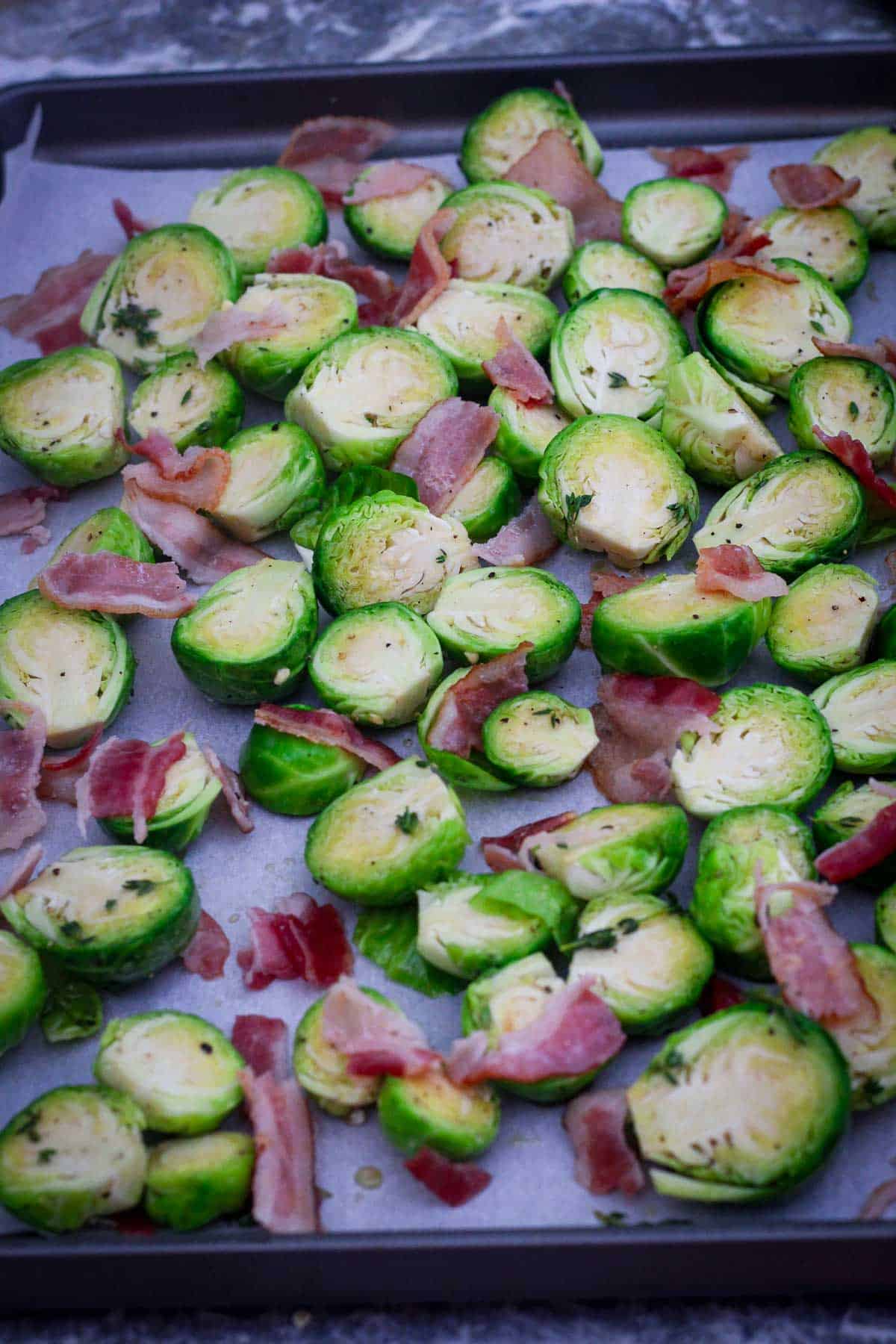 Marinated Brussel sprouts and bacon on a baking sheet lined up with parchment paper, ready to bake. 