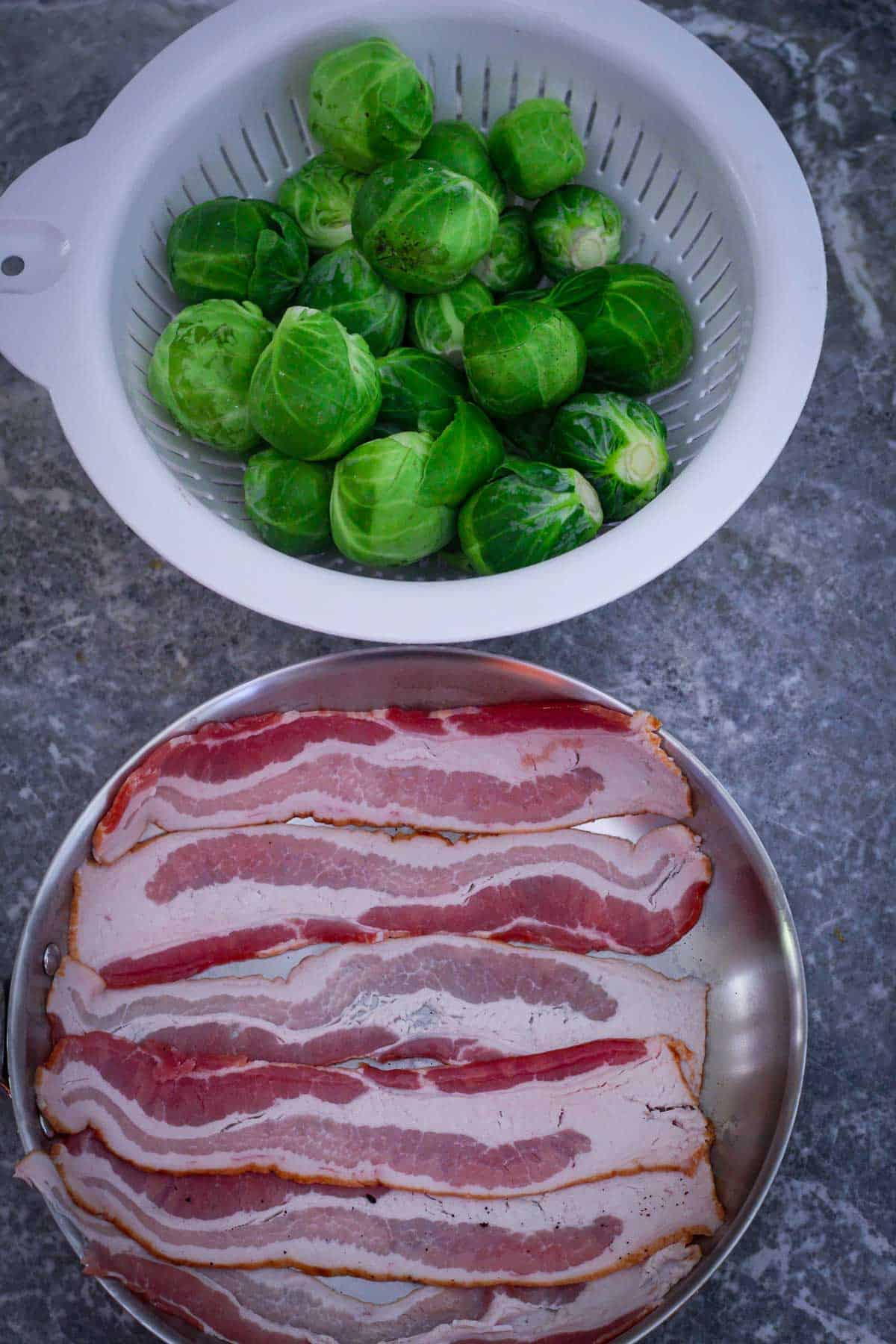A white colander full with brussel sprouts shown next to a pan with bacon strips. 