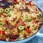 An oval Dutch oven showing a cooked chicken and rice dish. The food seems to have lots of veggies in the picture peaking through the rice like peppers, onions, olives etc.