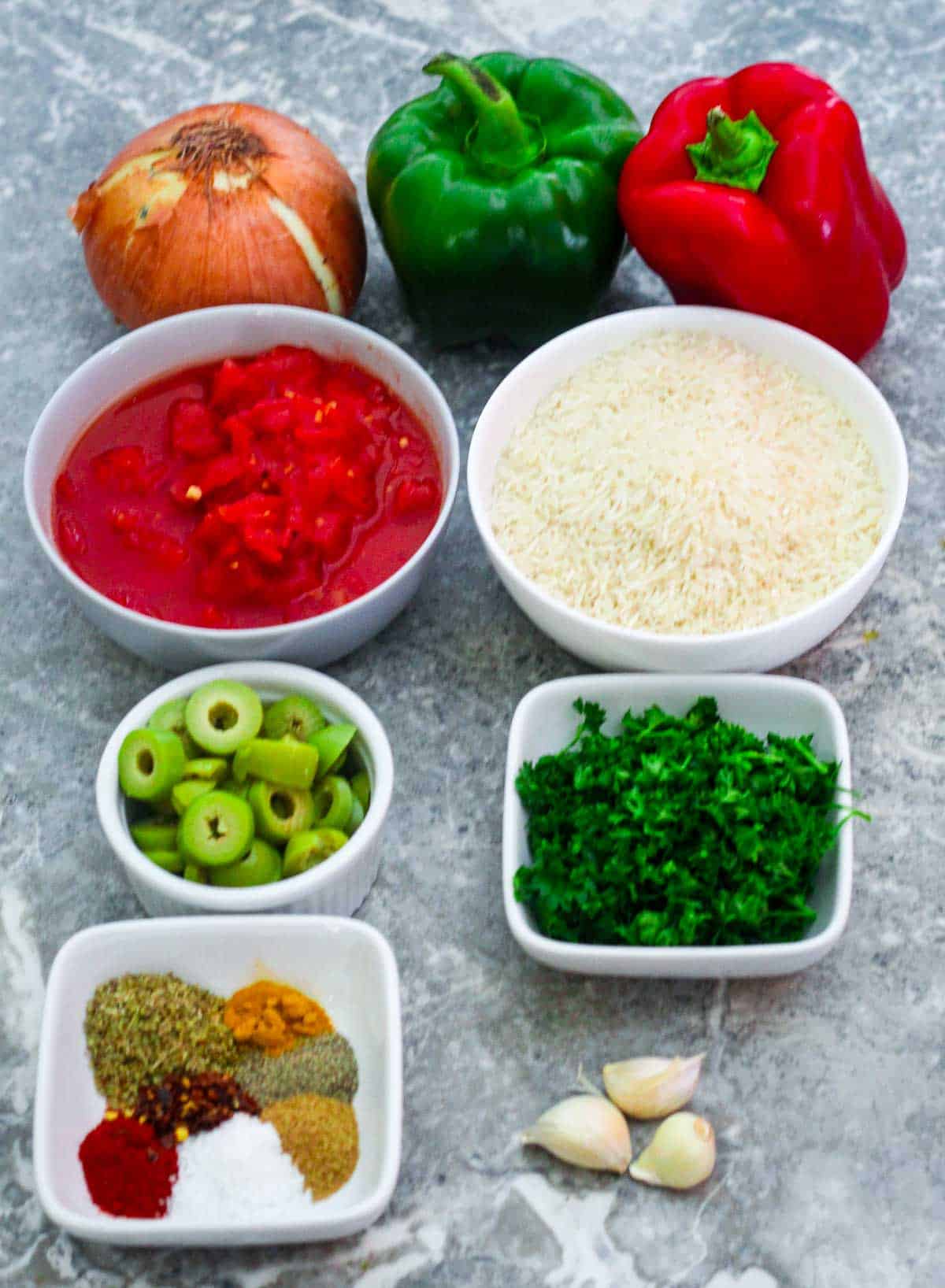 Some of the ingredients for the Spanish Chicken and Rice shown on a counter: onion, bell peppers, diced tomatoes, basmati rice, green olives, parsley, spices and garlic. 