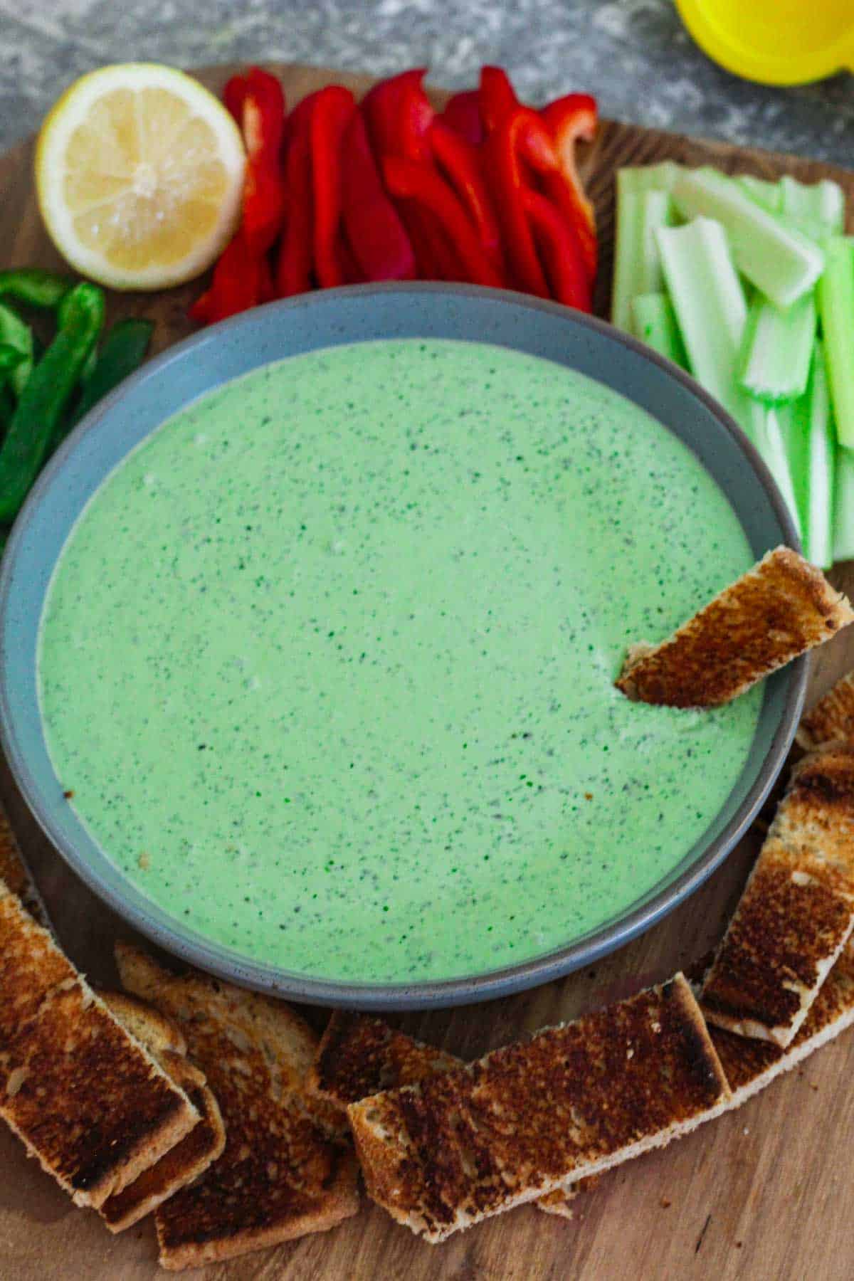 A green dip on a round plate, surrounded by veggies for dipping as well as strips of toasted bread. 