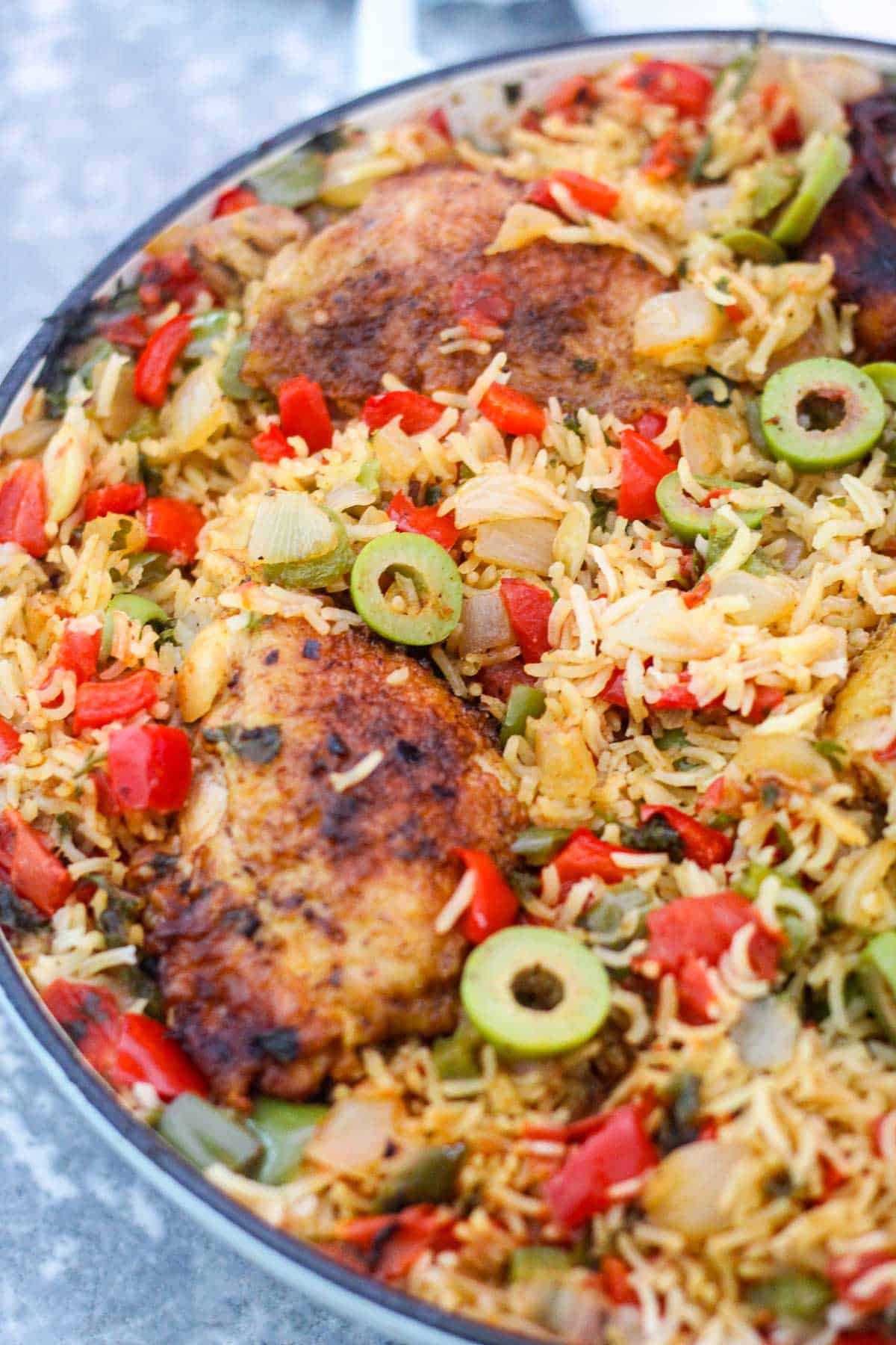 A pot showing cooked chicken, rice and lots of vegetables. Green olives, red peppers and onions are visible throughout the rice. 