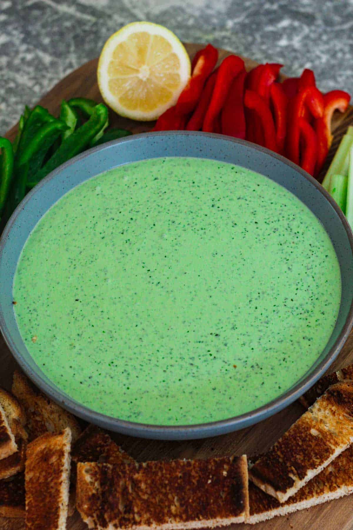 A green dip made with yogurt and herbs to create a healthy spin over the famous Green Goddess dressing. The green dip is shown in a plate, surrounded by red bell pepper strips, green bell peppers, celery, lemon, toasted sourdough bread etc. 