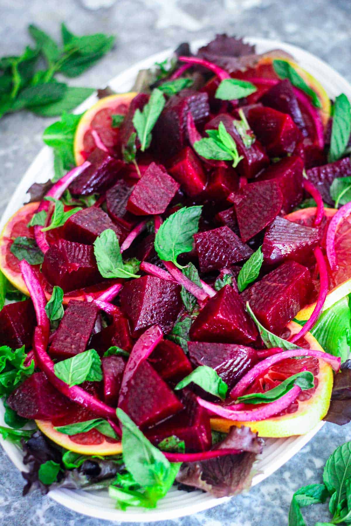 An oval platter with a beets and oranges salad over a bed of greens. You can see mint and onions peeking through other ingredients in the salad.