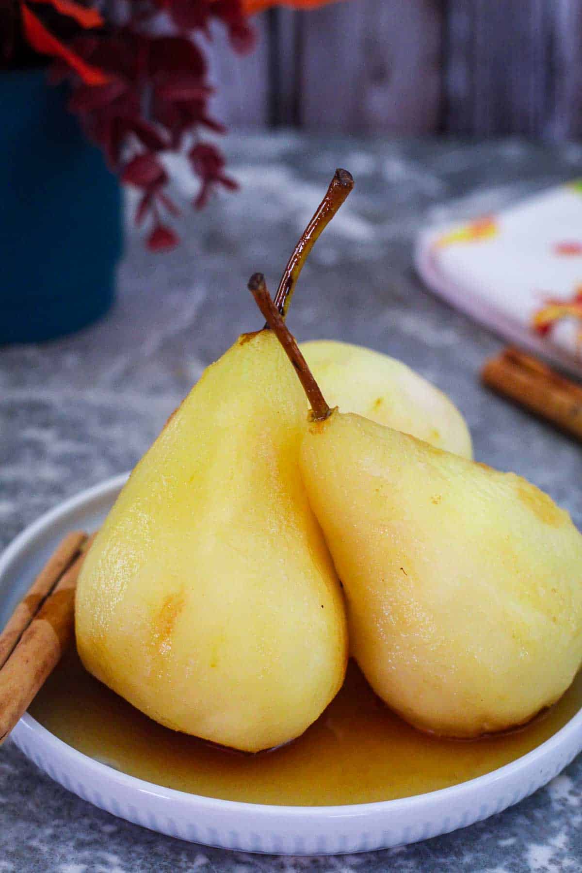 Poached pears served on a round plate, covered in syrup. They're garnished with a cinnamon stick.
