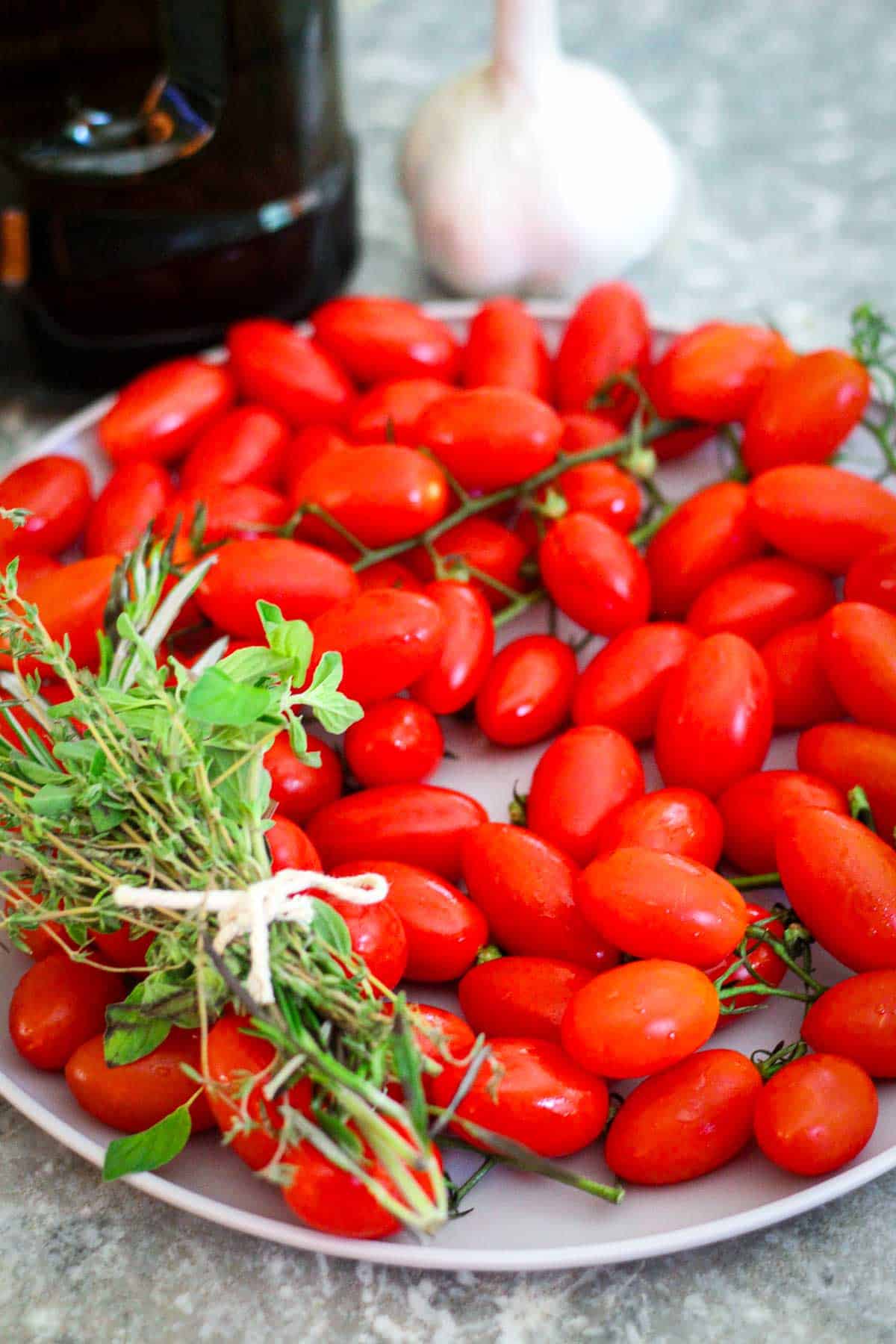 Fresh cherry tomatoes (some still in the vine), a bouquet of herbs tied with twine, garlic and oil are in the background.
