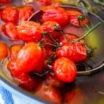 Tomatoes confit on a baking dish just finished cooking. There's a ladle bringing some of them still with stems on out of the oil for better view.
