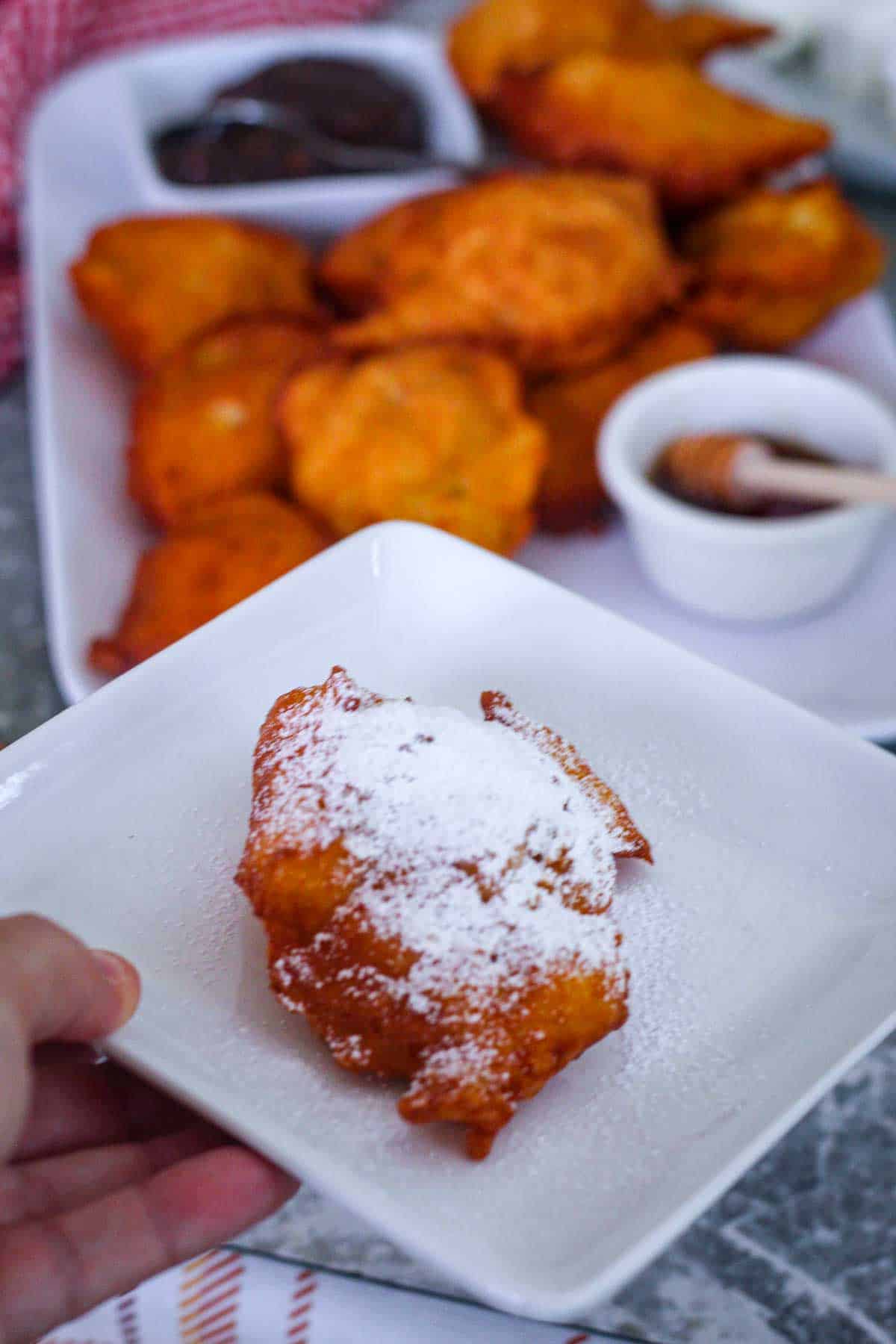 One fried dough (petulla) covered in powder sugar on a plate with the rest of fried petulla on a platter in the background.