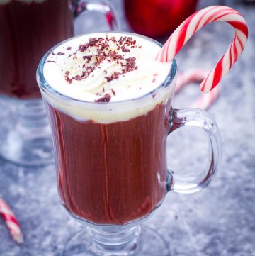 Hot chocolate in a glass cup, topped with whipped cream, chocolate and candy cane.