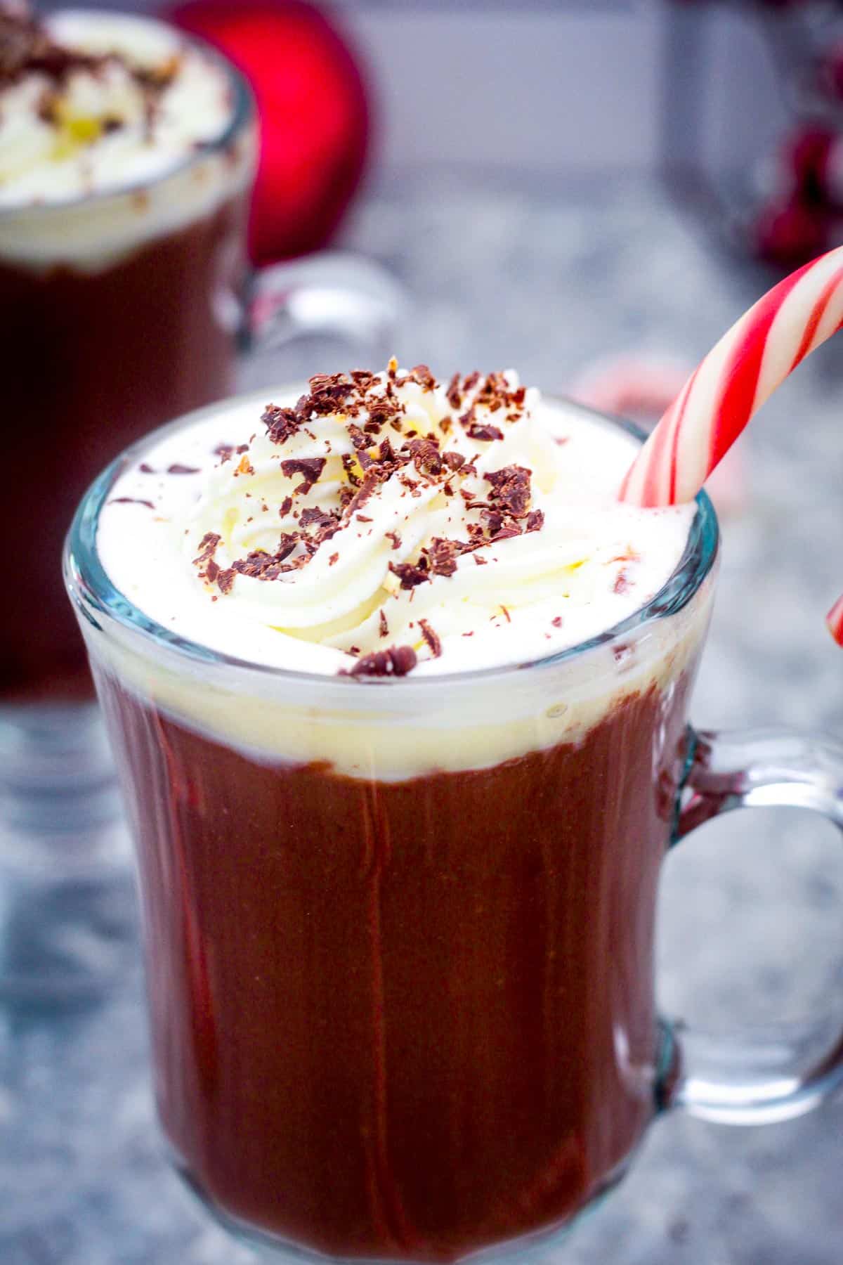 A glass cup of hot chocolate with whipped cream, shaved chocolate over the cream and a candy cane dipped on the chocolate. 