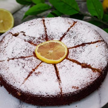 Round lemon ricotta cake, sliced in 8 and has a slice of lemon in the middle of the cake. Cake is dusted with powder sugar.