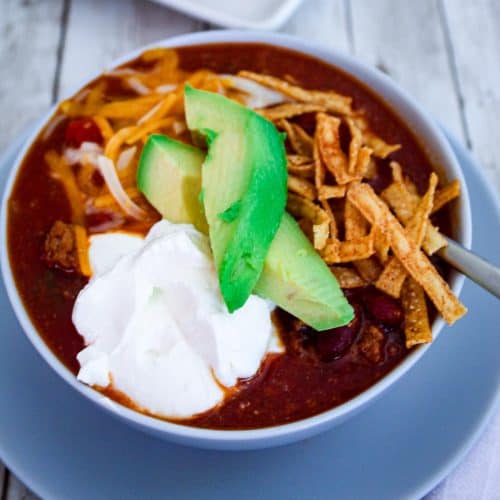 A bowl of chili topped with sour cream, avocado slices, tortilla strips, cheddar cheese.