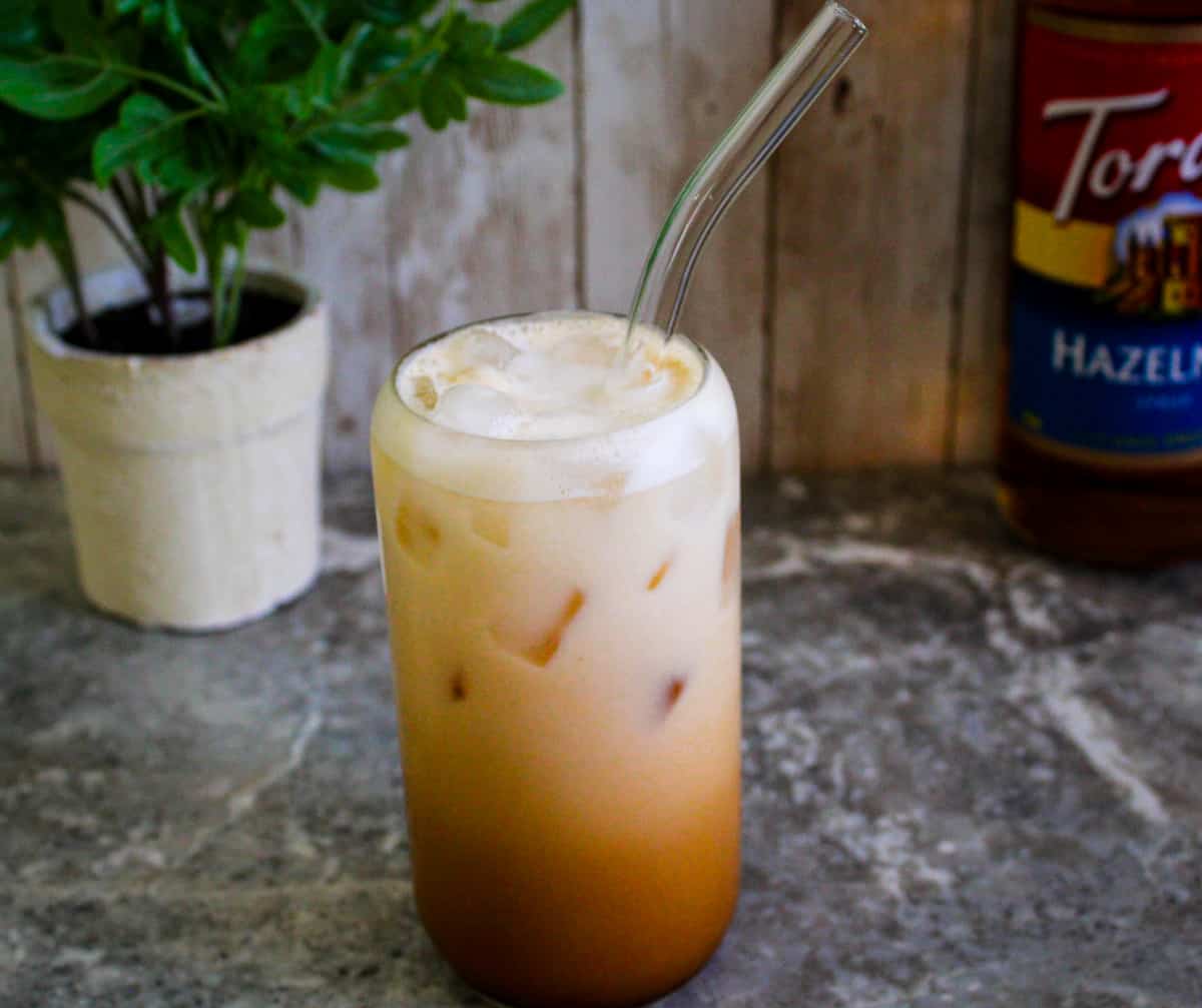 Shaken espresso drink with ice and hazelnut syrup served on a glass cup with a glass straw.