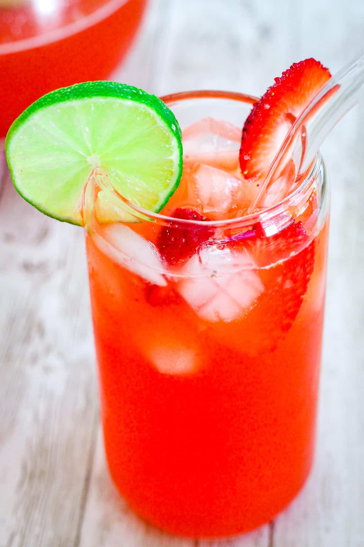 A glass with strawberry limeade drink, it's shown with a lime slice and strawberry slice as garnishes. Drink has pieces of fruit and ice in it.