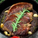 A steak cooked in cast iron, shown in a close up shot. Steak is garnished with rosemary, and there's more herbs and garlic around it in the skillet.