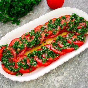 A tomato salad platter with sliced tomatoes and a parsley marinade on top.